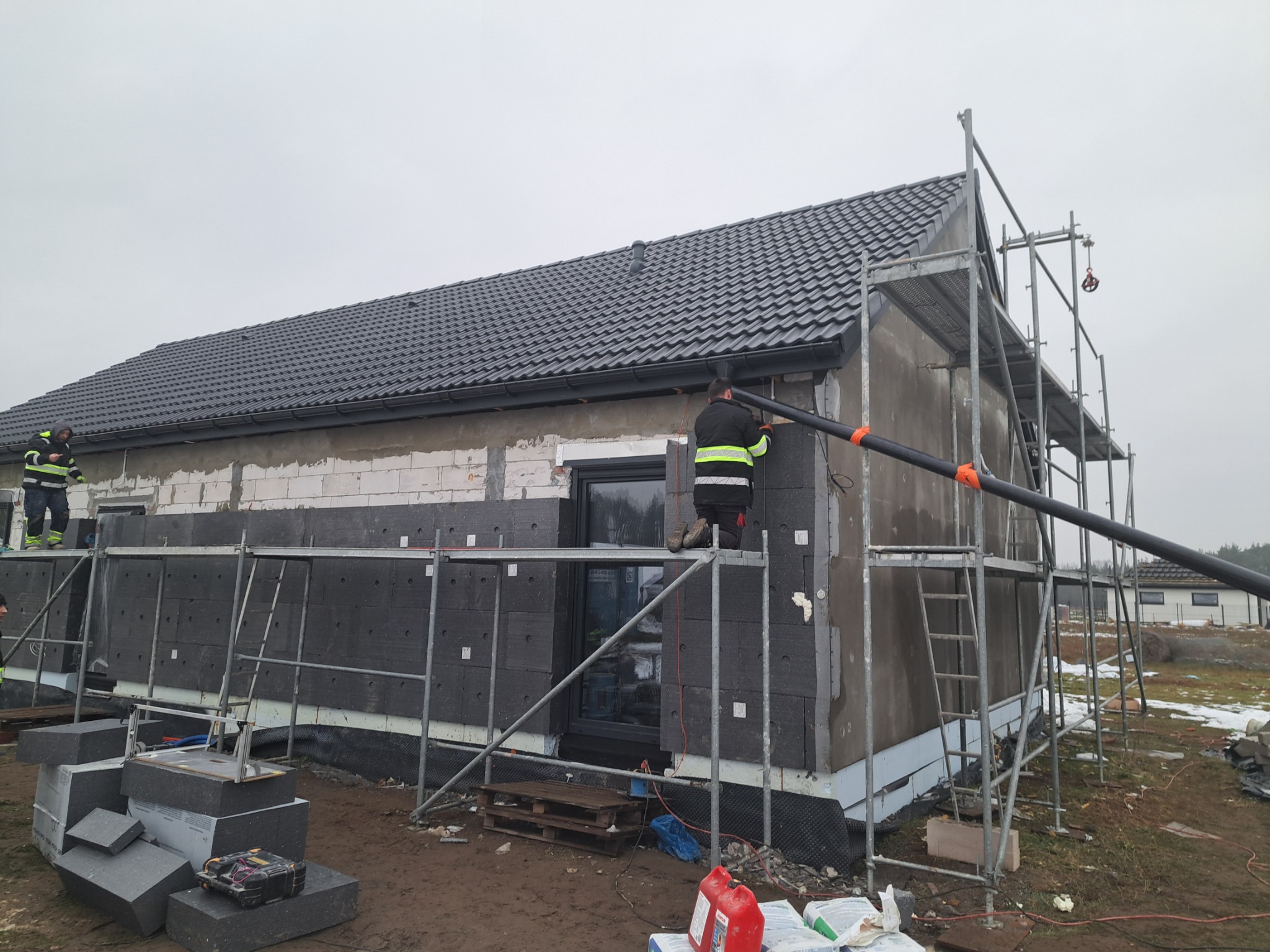 Ocieplanie domu styropianem grafitowym na rusztowaniu. Widoczni dwaj pracownicy w odblaskowych ubraniach. Na ziemi leżą płyty styropianowe i narzędzia.