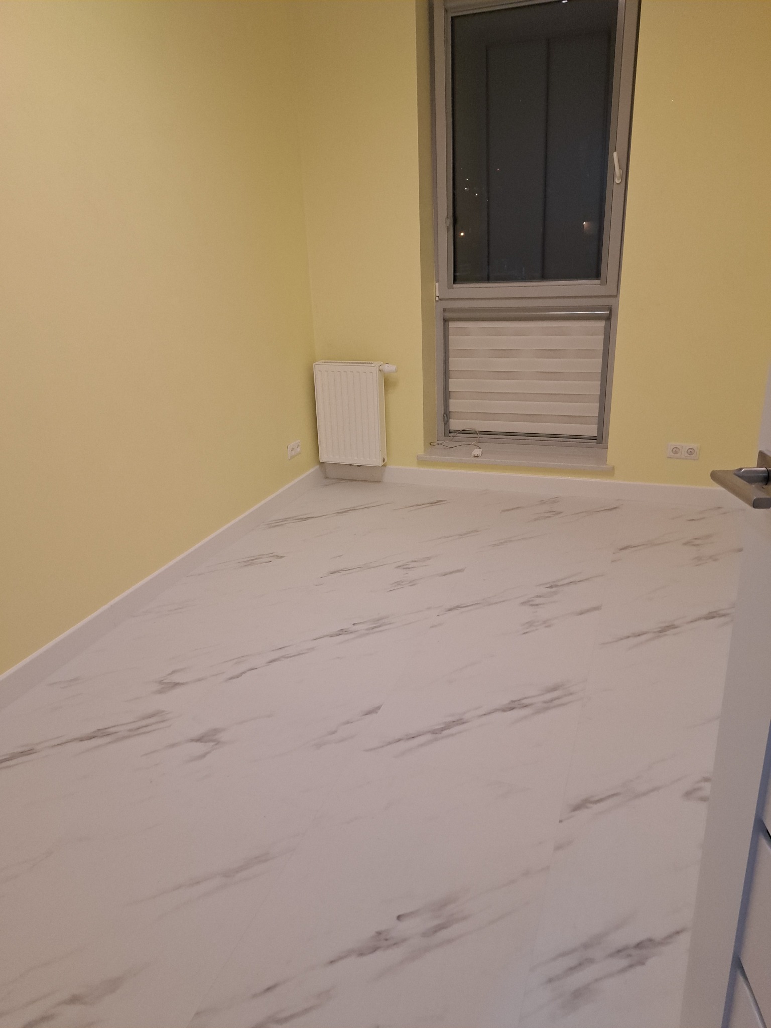 Pomalowane na żółto wnętrze pokoju z marmurową podłogą, grzejnikiem i oknem z żaluzjami. Widoczne gniazdka elektryczne i fragment drzwi z klamką.