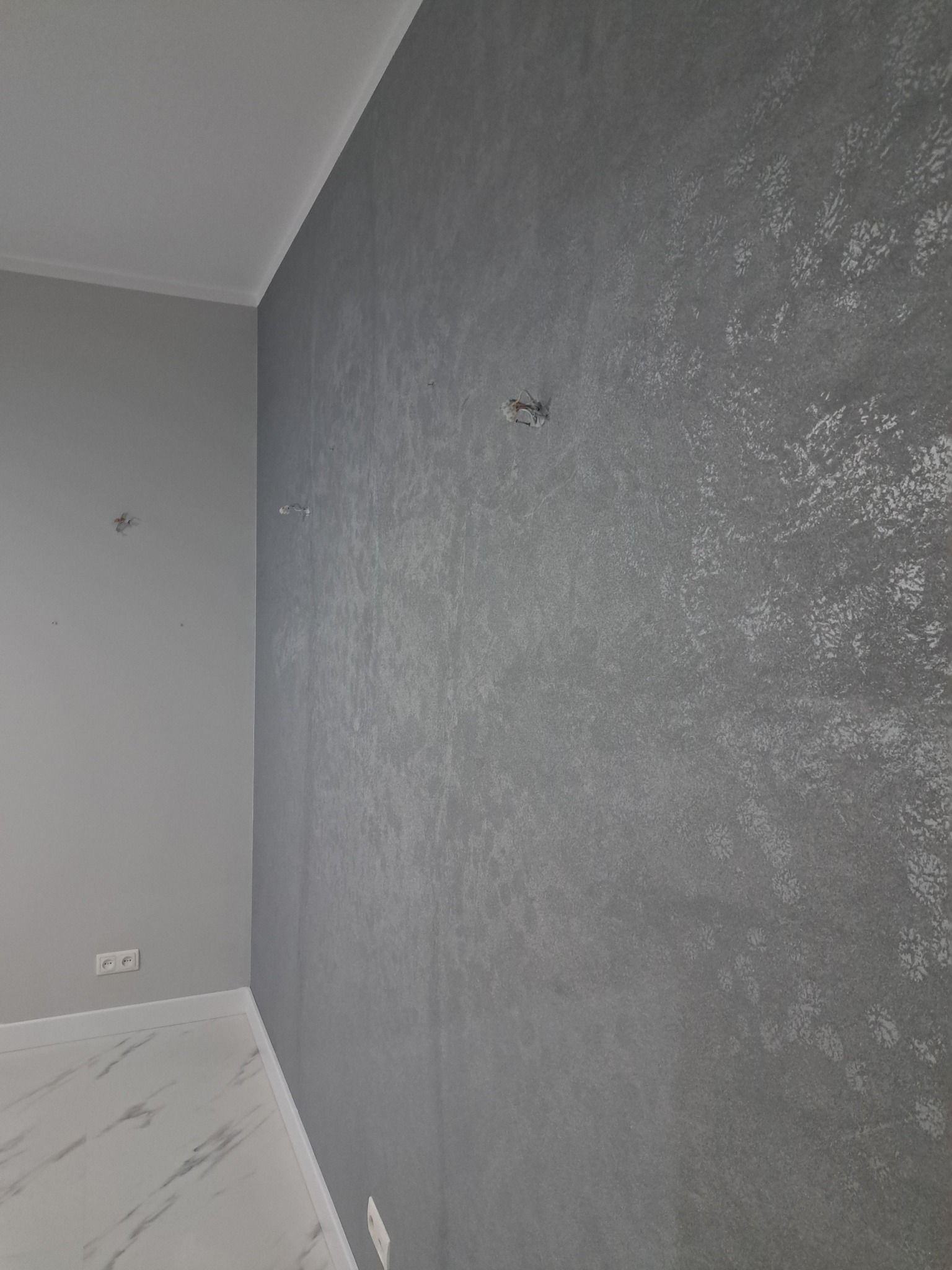 Szara ściana z dekoracyjną fakturą w nowym wnętrzu. Widoczne gniazdka elektryczne i marmurowa podłoga. Minimalistyczny design.