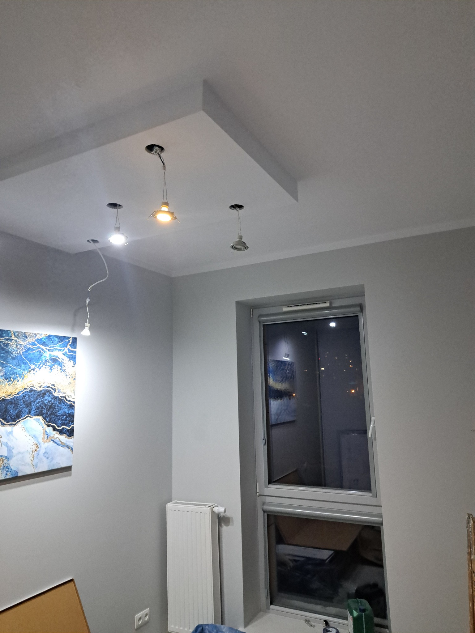 Szare wnętrze z oknem i trzema lampami sufitowymi, z których jedna świeci. Na ścianie wisi obraz w niebieskich tonacjach. Widoczny grzejnik.