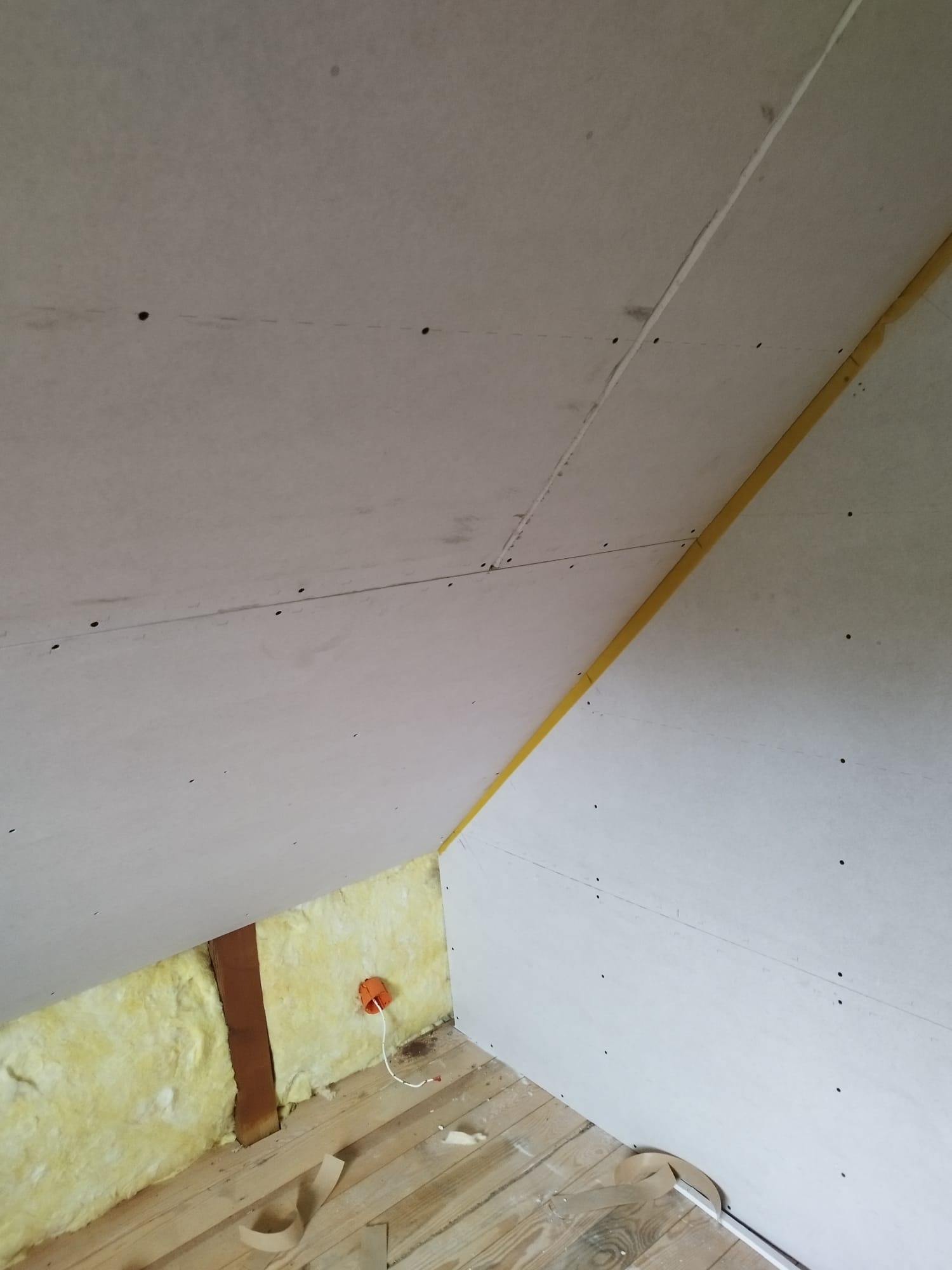 Skośny sufit poddasza z płyt karton-gips, widoczne łączenia wzmocnione taśmą, ocieplenie z wełny mineralnej, drewniana podłoga i instalacja elektryczna.