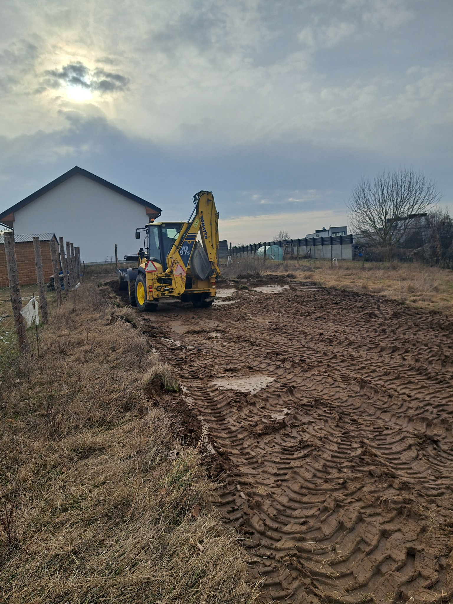 Żółta koparko-ładowarka Komatsu WB97S na błotnistym terenie, ślady opon, w tle dom i pochmurne niebo. Przygotowanie terenu pod budowę.