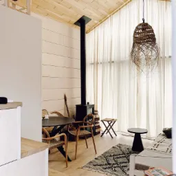 Jasne wnętrze z drewnianym sufitem, piecem wolnostojącym i wysokimi, kremowymi zasłonami przy oknie, oświetlone wiszącą lampą z naturalnych materiałów.