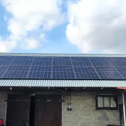 Instalacja paneli słonecznych na dachu budynku gospodarczego z blachy falistej, widoczne niebo z chmurami.