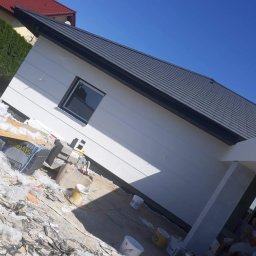 MARTOS Usługi budowlane Mariusz Gaca - Świeżo otynkowana elewacja domu jednorodzinnego z widocznymi śladami prac budowlanych, styropianem i wiadrami po farbie na ziemi, czyste niebo w tle.