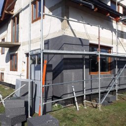 MARTOS Usługi budowlane Mariusz Gaca - Ocieplanie ściany zewnętrznej domu styropianem grafitowym, widoczne rusztowanie, poziomica i stos płyt izolacyjnych na trawie.