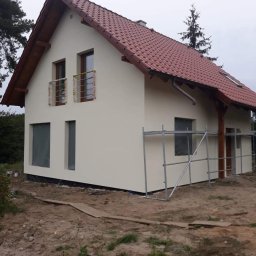 MARTOS Usługi budowlane Mariusz Gaca - Dom jednorodzinny z jasną elewacją, częściowo zakryte okna folią ochronną, rusztowanie przy ścianie, czerwona dachówka, teren wokół w stanie przygotowania.