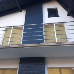 MARTOS Usługi budowlane Mariusz Gaca - Fragment elewacji domu z balkonem, okna zabezpieczone folią malarską, pionowy pas elewacji wykończony ciemną cegłą klinkierową, biała ściana, grafitowy dach.