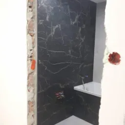 Częściowo wykończona łazienka z czarnymi płytkami imitującymi marmur na ścianie, białą wanną i surowym otworem drzwiowym z widoczną cegłą i instalacją elektryczną.