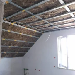 Poddasze w trakcie ocieplania wełną mineralną, widoczne metalowe profile konstrukcji pod płytę kartonowo-gipsową, nieotynkowane ściany z instalacją hydrauliczną i elektryczną.