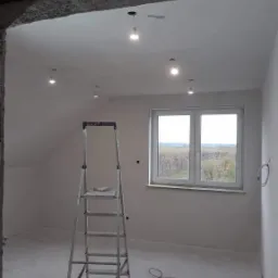 Wnętrze pokoju po remoncie: gładkie, białe ściany, sufit z punktowymi lampami, okno z widokiem na drzewa, aluminiowa drabina z narzędziami na podeście i czerwony przewód elektryczny na podłodze.