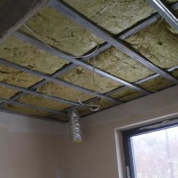 MARTOS Usługi budowlane Mariusz Gaca - Sufit z metalową konstrukcją wypełnioną żółtą wełną mineralną, widoczny fragment wentylacji w folii aluminiowej zwisający z sufitu, okno w tle.