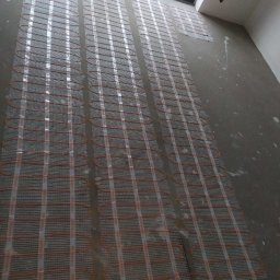 MARTOS Usługi budowlane Mariusz Gaca - Rozłożona siatka grzewcza z pomarańczowymi kablami, przygotowana do zalania wylewką, widok z góry, surowy stan wykończenia, duże okno w tle.