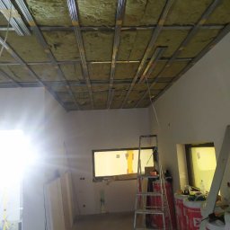 MARTOS Usługi budowlane Mariusz Gaca - Montaż izolacji z wełny mineralnej i konstrukcji metalowej pod płytę karton gips na suficie w trakcie remontu, widoczna drabina i materiały budowlane.