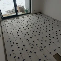 Jasne panele podłogowe ułożone w pokoju, z użyciem klipsów dystansowych, widok na okno z zimowym krajobrazem na zewnątrz, surowa żarówka zwisająca z sufitu.