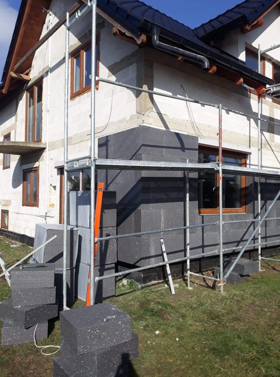 Ocieplanie ściany zewnętrznej domu styropianem grafitowym, widoczne rusztowanie, poziomica i stos płyt izolacyjnych na trawie.