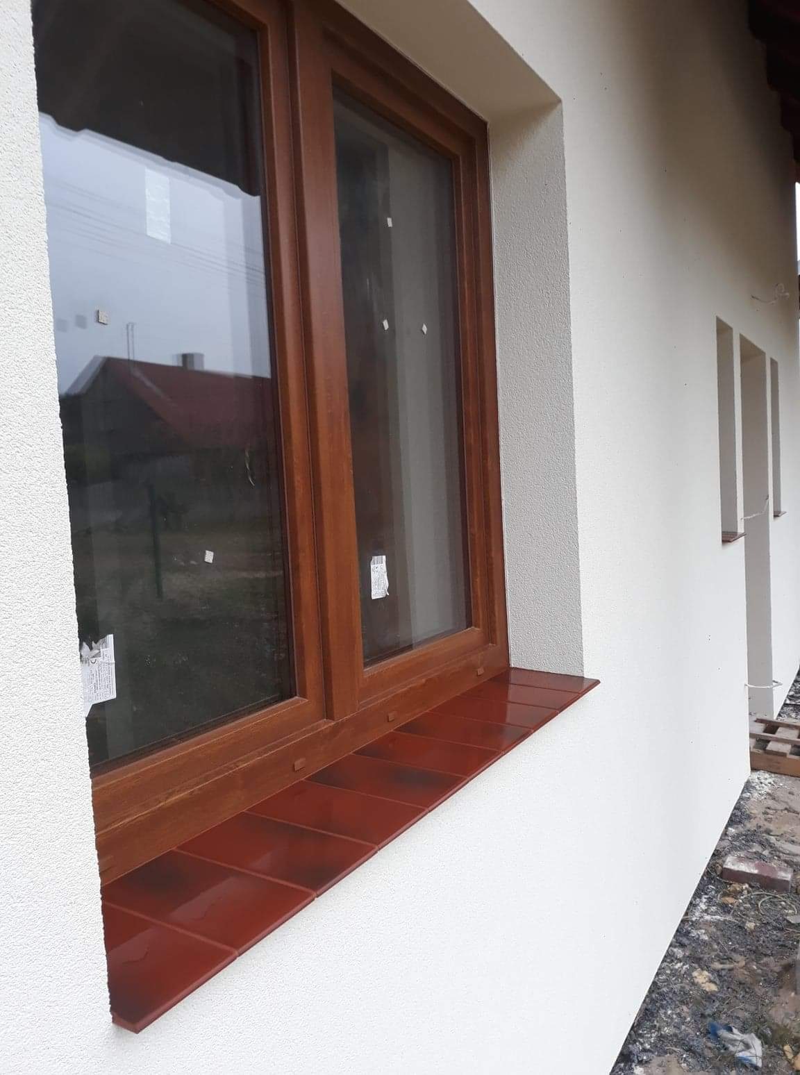 Nowe, brązowe okno PCV z parapetem wykończonym klinkierem, osadzone w świeżo otynkowanej, białej elewacji budynku.