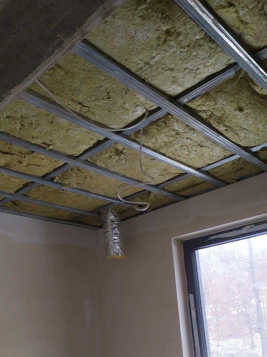 Sufit z metalową konstrukcją wypełnioną żółtą wełną mineralną, widoczny fragment wentylacji w folii aluminiowej zwisający z sufitu, okno w tle.