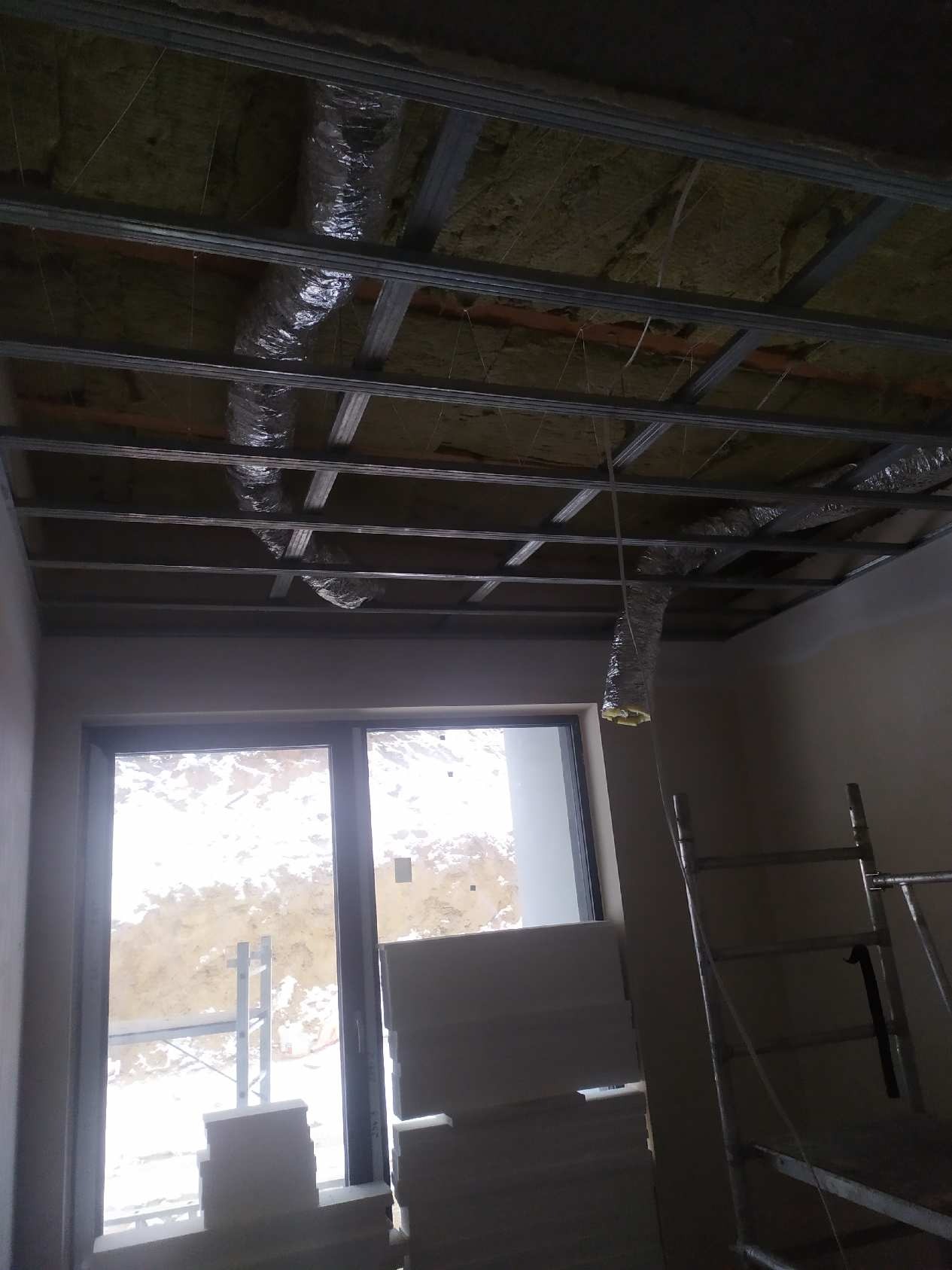 Sufit w trakcie montażu stelaża z profili metalowych i izolacji z wełny mineralnej, widoczne przewody wentylacyjne w folii aluminiowej, okno i drabina w tle.