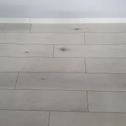 Ułożona jasna podłoga panelowa z widocznymi słojami drewna, przy białej listwie przypodłogowej i białej ścianie.