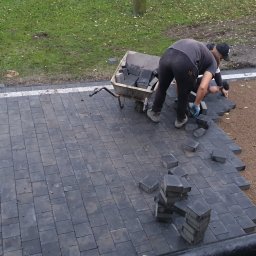 Wieliczka - Układanie ciemnoszarej kostki brukowej z białym obramowaniem, widoczni dwaj pracownicy w trakcie pracy, taczka z kostką i łopata w tle.
