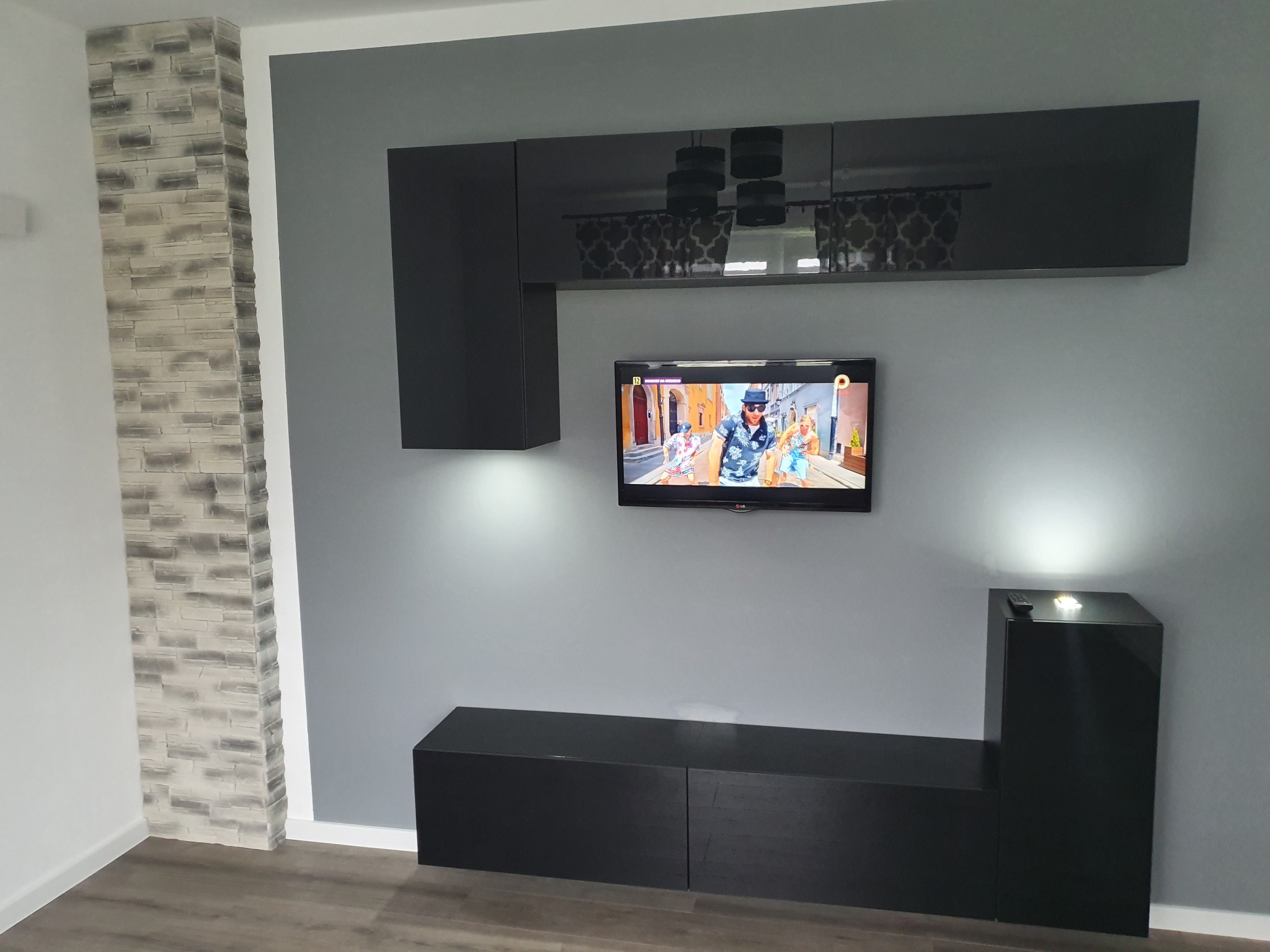 Szara ściana z czarnymi, błyszczącymi szafkami wiszącymi i stojącymi, telewizor na ścianie, dekoracyjny panel imitujący kamień na ścianie po lewej stronie.