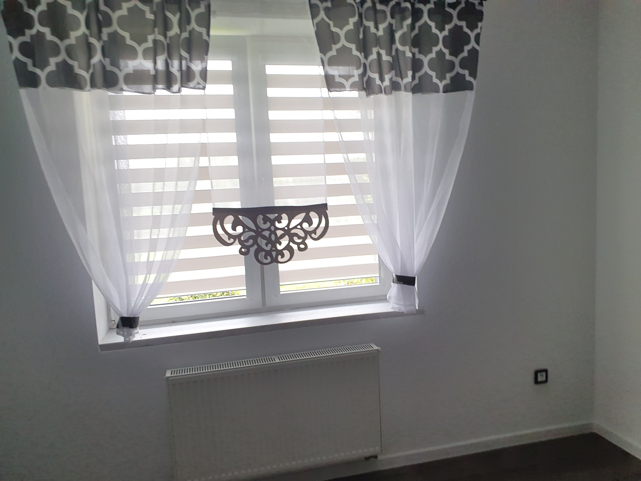 Okno z żaluzjami poziomymi i dekoracyjnymi zasłonami w odcieniach szarości, ozdobny element dekoracyjny na szybie, grzejnik pod oknem.