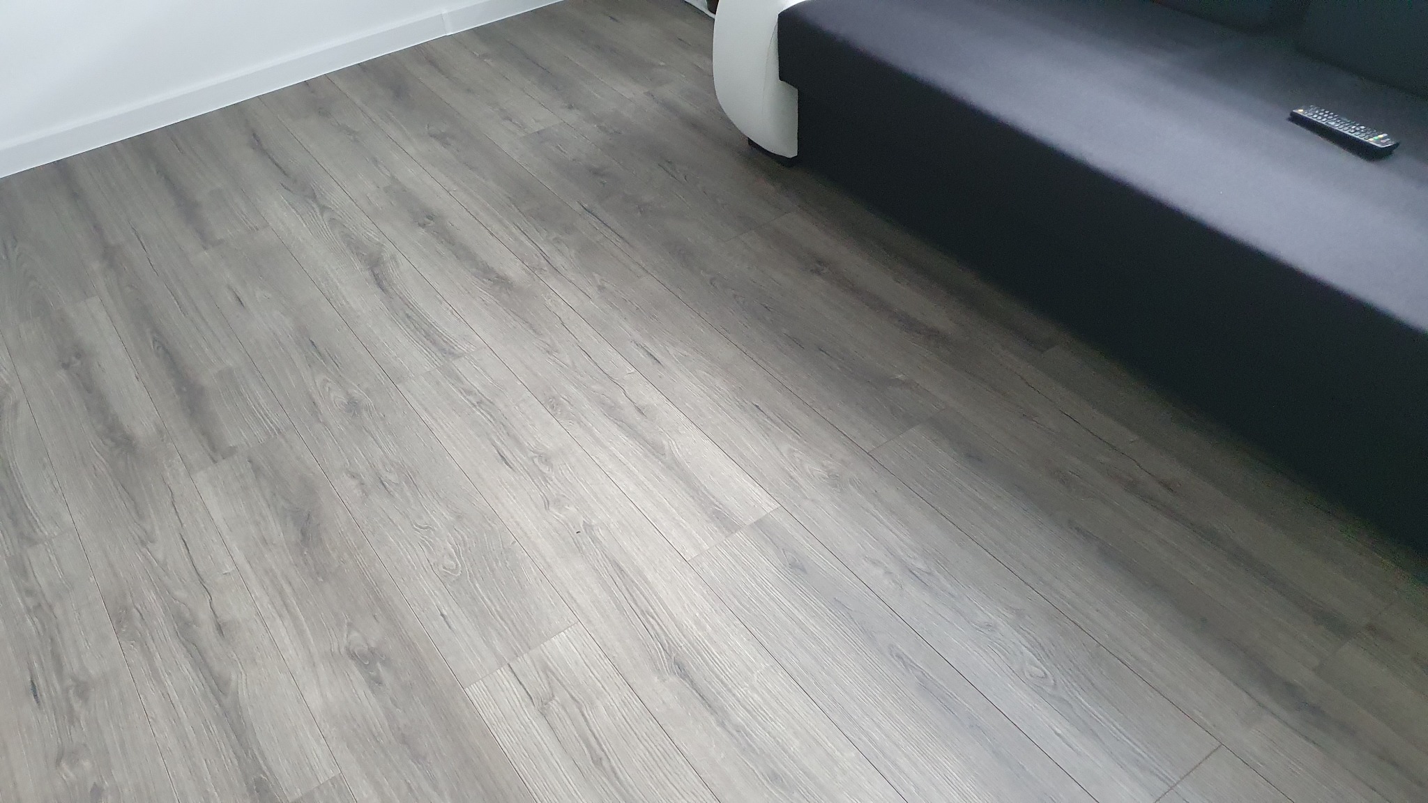 Szara podłoga z paneli imitujących drewno, ułożona ukośnie, widoczny fragment sofy i pilota.