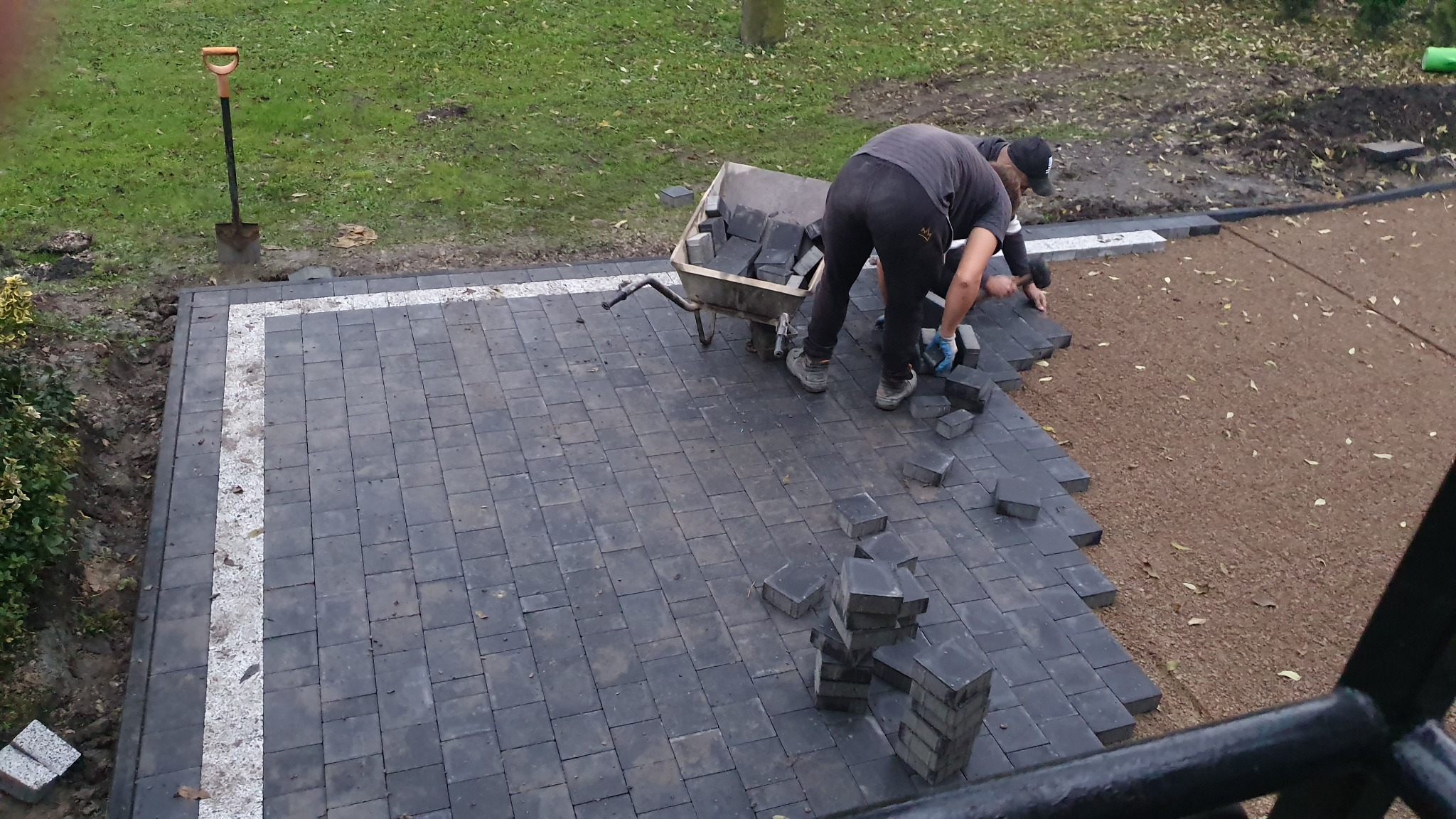 Układanie ciemnoszarej kostki brukowej z białym obramowaniem, widoczni dwaj pracownicy w trakcie pracy, taczka z kostką i łopata w tle.