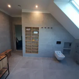 Szara łazienka na poddaszu z oknem dachowym, wyposażona w bidet, grzejnik drabinkowy, wnękę z półkami i drewniany regał w ścianie.