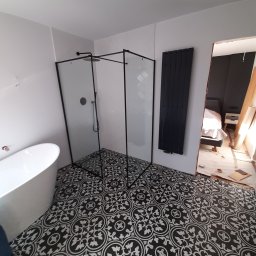 LUXPUR PAUL KUŚNIERZ-GAZDA - Nowoczesna łazienka w trakcie remontu: wolnostojąca wanna, kabina prysznicowa z czarnymi ramami, dekoracyjna czarno-biała podłoga, grzejnik drabinkowy, widok na sypialnię w tle