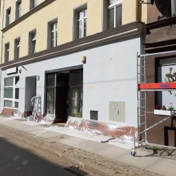 LUXPUR PAUL KUŚNIERZ-GAZDA - Prace malarskie na elewacji budynku usługowego, widoczne rusztowanie i zabezpieczona folią strefa wejścia, pracownik na drabinie przy oknie.