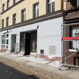 Prace malarskie na elewacji budynku usługowego, widoczne rusztowanie i zabezpieczona folią strefa wejścia, pracownik na drabinie przy oknie.