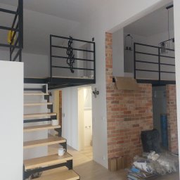 LUXPUR PAUL KUŚNIERZ-GAZDA - Wnętrze mieszkania z antresolą, schodami z drewnianymi stopniami na metalowej konstrukcji, fragmentem ściany z czerwonej cegły, widoczne elementy oświetlenia i kubek na schodach.