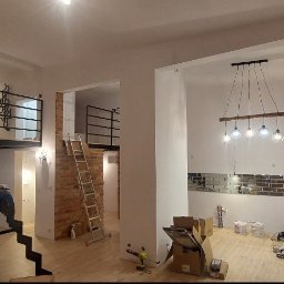 LUXPUR PAUL KUŚNIERZ-GAZDA - Wnętrze mieszkania w trakcie prac wykończeniowych: widoczne schody z metalową balustradą prowadzące na antresolę, ceglana ściana z drabiną, zamontowane oświetlenie i lustrzane płytki na ścianie...