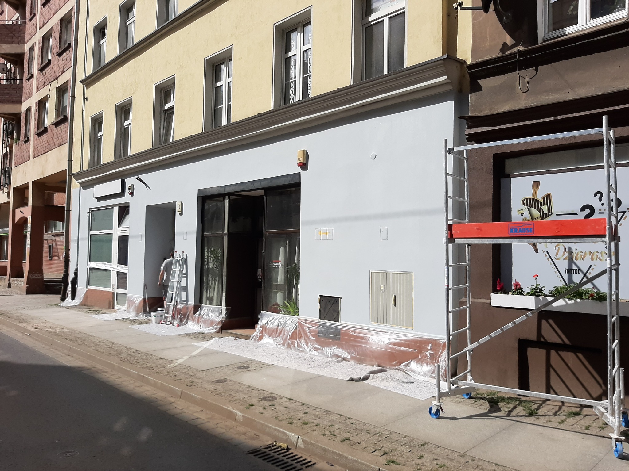 Prace malarskie na elewacji budynku usługowego, widoczne rusztowanie i zabezpieczona folią strefa wejścia, pracownik na drabinie przy oknie.
