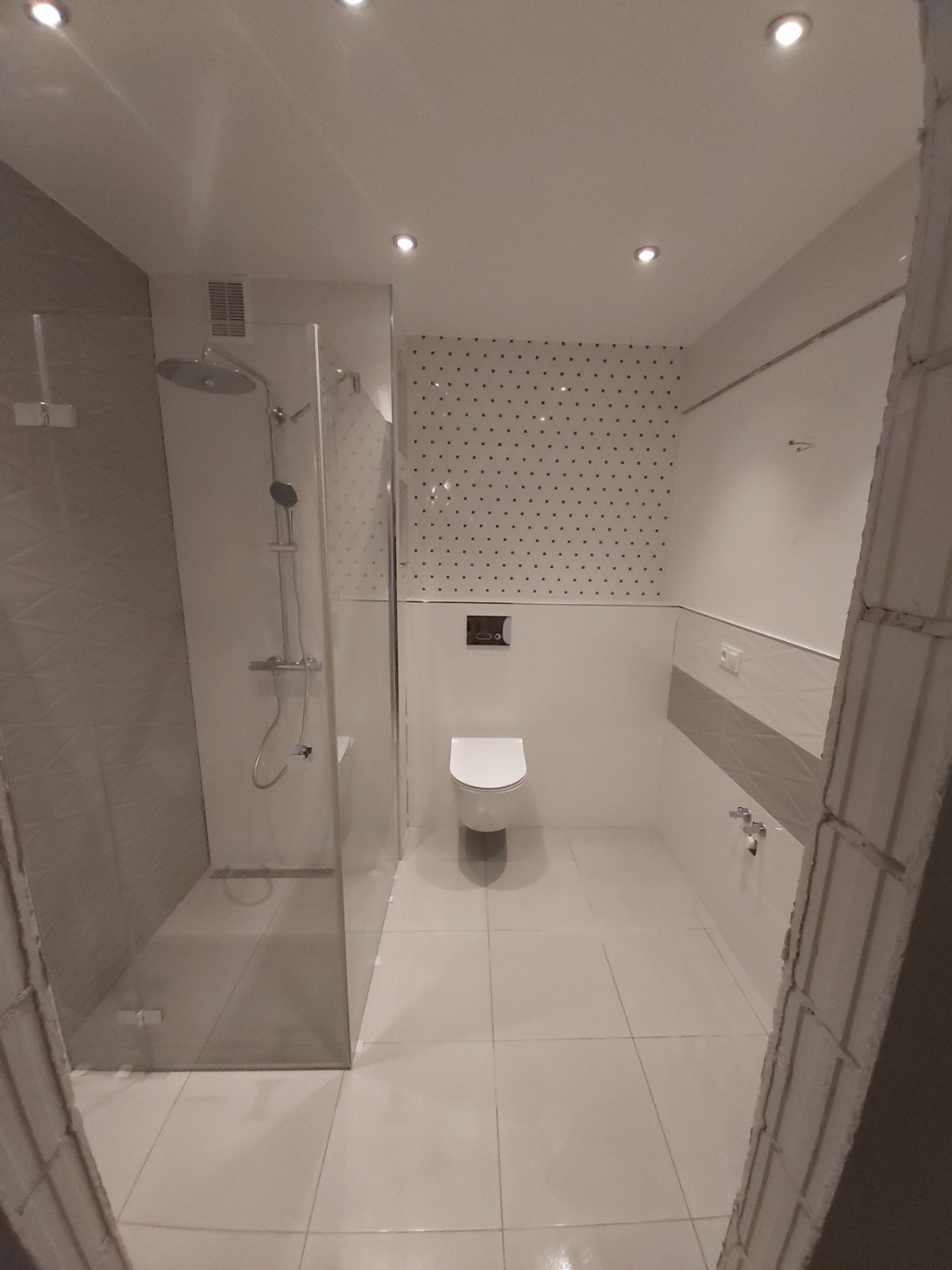 Nowoczesna łazienka z kabiną prysznicową typu walk-in, toaletą podwieszaną i ścianą w czarne kropki, wykończona w jasnej kolorystyce.