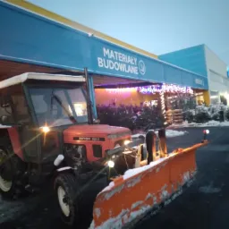 Czerwony traktor Zetor 5211 z pomarańczowym pługiem do śniegu stoi na parkingu przed sklepem budowlanym o zmierzchu. Pług pokryty śniegiem, w tle oświetlone drzewka świąteczne.