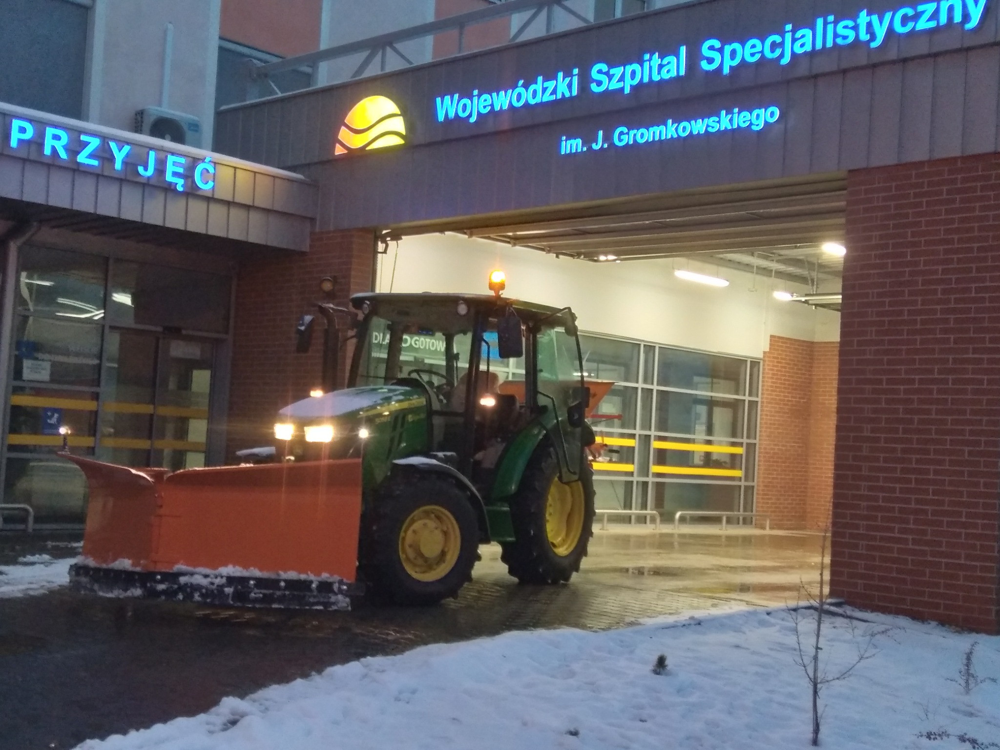 Zielony traktor z pomarańczowym pługiem odśnieżającym przed budynkiem Wojewódzkiego Szpitala Specjalistycznego we Wrocławiu, na ziemi resztki śniegu.