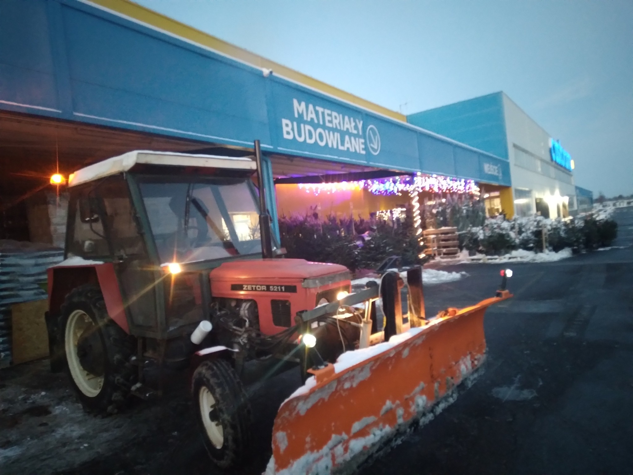Czerwony traktor Zetor 5211 z pomarańczowym pługiem do śniegu stoi na parkingu przed sklepem budowlanym o zmierzchu. Pług pokryty śniegiem, w tle oświetlone drzewka świąteczne.