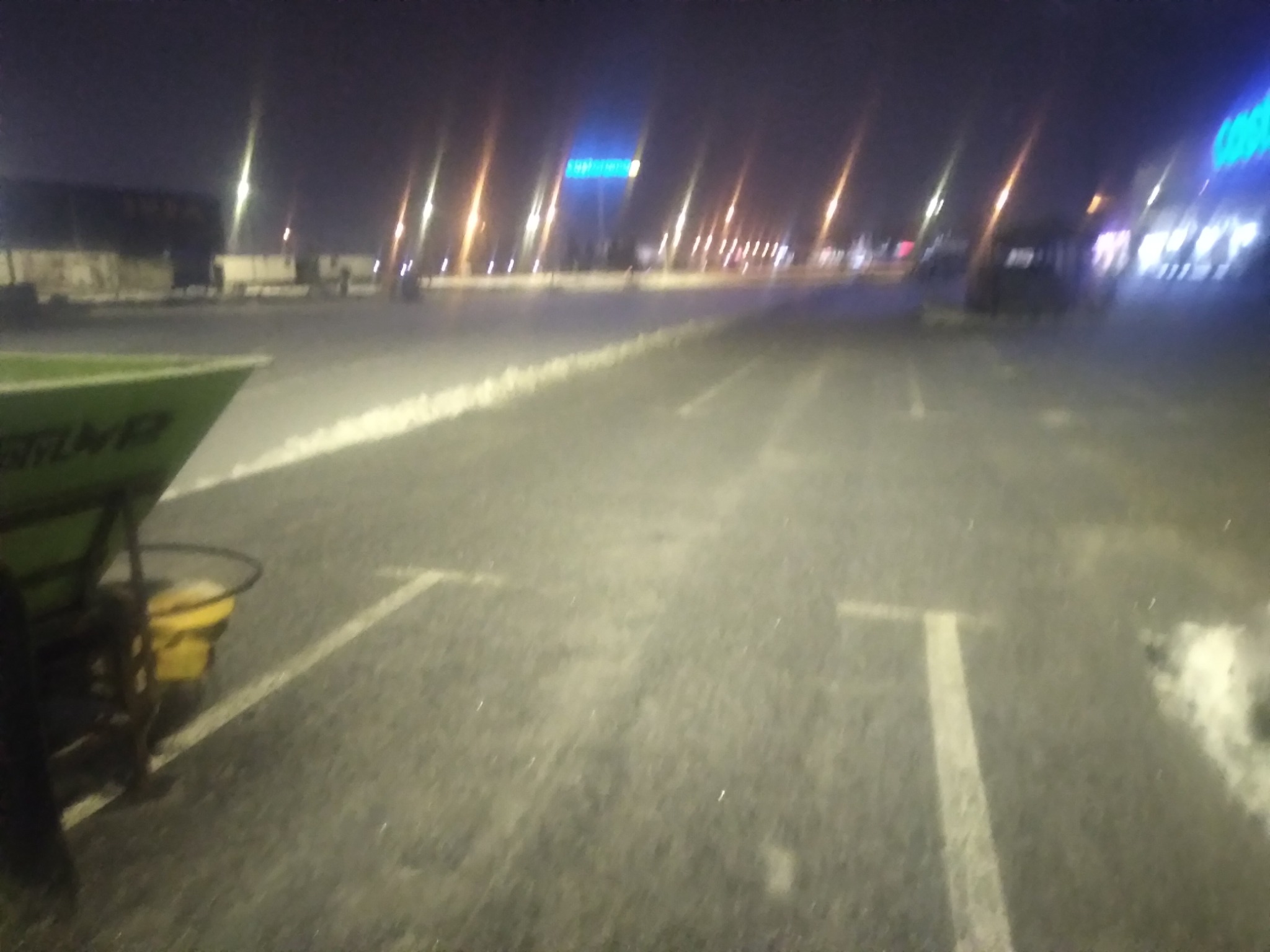 Nocne ujęcie parkingu po odśnieżaniu, widoczny rozsypywacz soli na wózku, białe linie wyznaczające miejsca parkingowe, oświetlone budynki w tle.