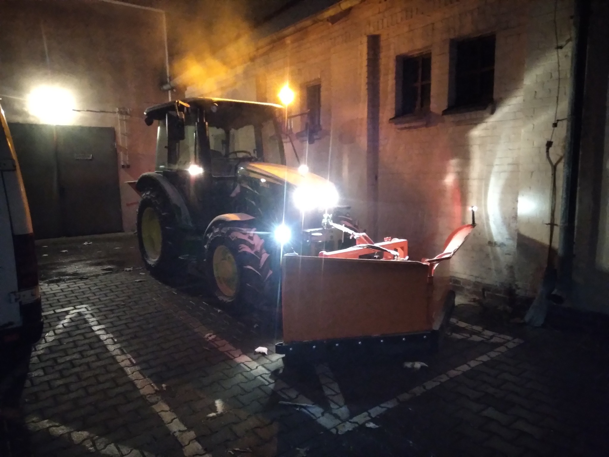 Ciągnik John Deere z pługiem śnieżnym, oświetlony reflektorami, na brukowanym placu przy budynku z cegły w nocy.