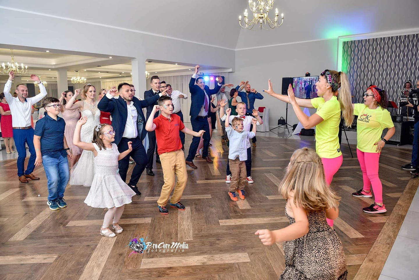 Grupa ludzi, w tym dzieci z pomalowanymi twarzami, tańcząca na parkiecie podczas uroczystości z dwoma animatorkami w jaskrawych strojach, prowadzącymi zabawę.