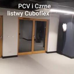 Jasna podłoga z PCV i czarne listwy przypodłogowe Cuboflex w nowym wnętrzu, widok na szklane drzwi i grzejnik na ciemnej ścianie.