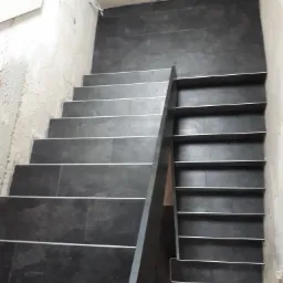 Schody wyłożone ciemnymi płytkami z metalowymi listwami, ściany w stanie surowym, widok z góry.