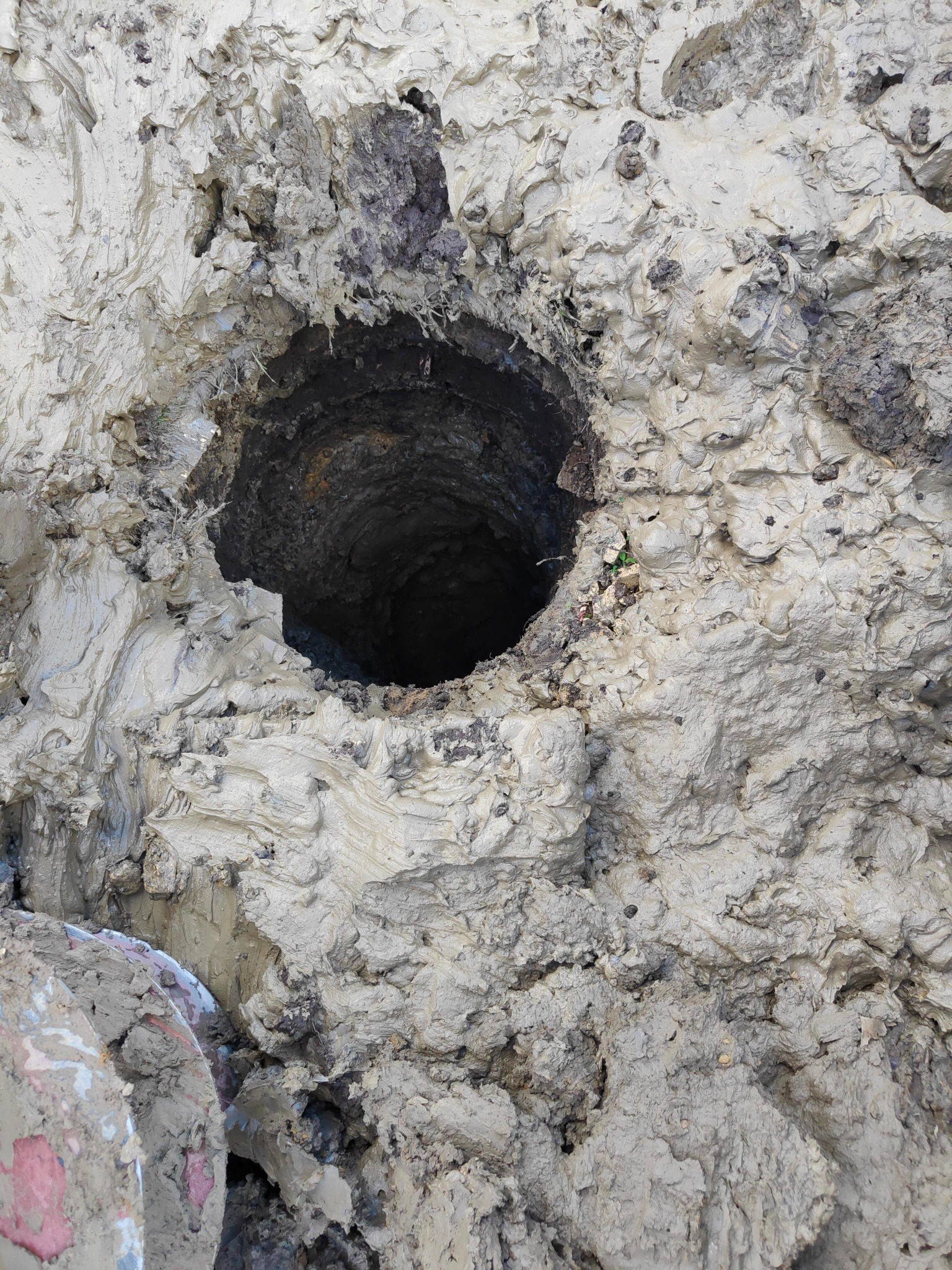 Wywiercony otwór w mokrej, gliniastej ziemi, widoczne ślady wiercenia i resztki ziemi na krawędziach otworu, fragment rury w lewym dolnym rogu.