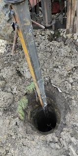 Wiertnica zagłębiająca się w ziemię, widok z góry na otwór w ziemi z wystającą wiertarką, fragmenty trawy i błota wokół, sprzęt budowlany w tle.