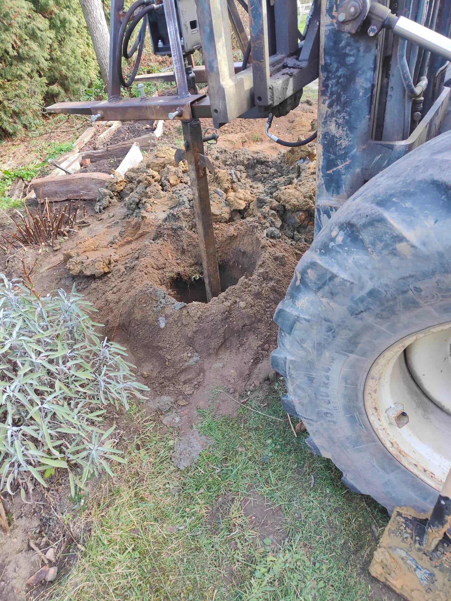 Wiertnica zagłębia się w ziemię, przygotowując otwór pod instalację, z widocznym fragmentem opony maszyny i zielenią w tle.