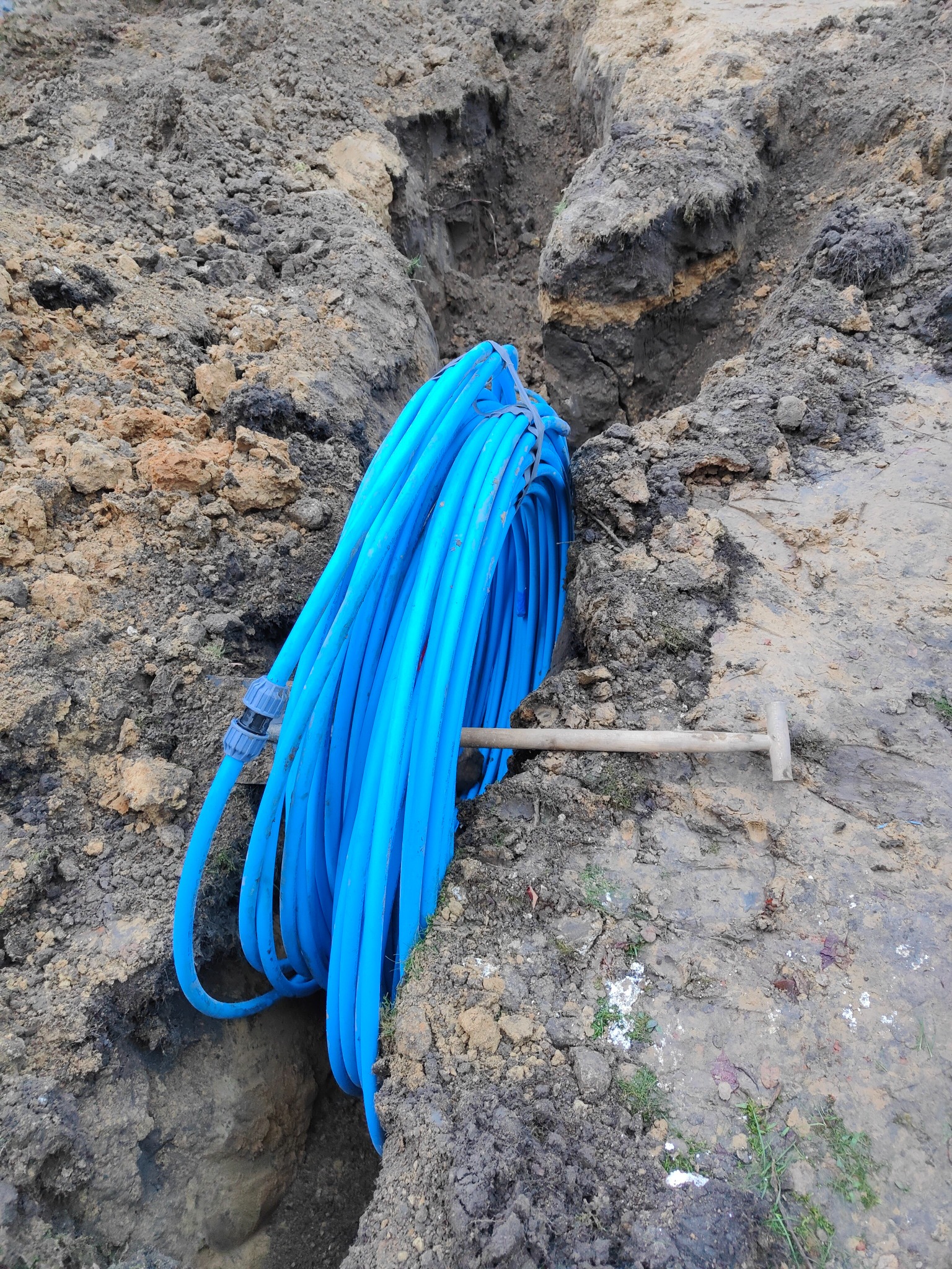 Zwój niebieskiego węża złączonego złączką leży obok wykopu i wbitego szpadla w ziemi.