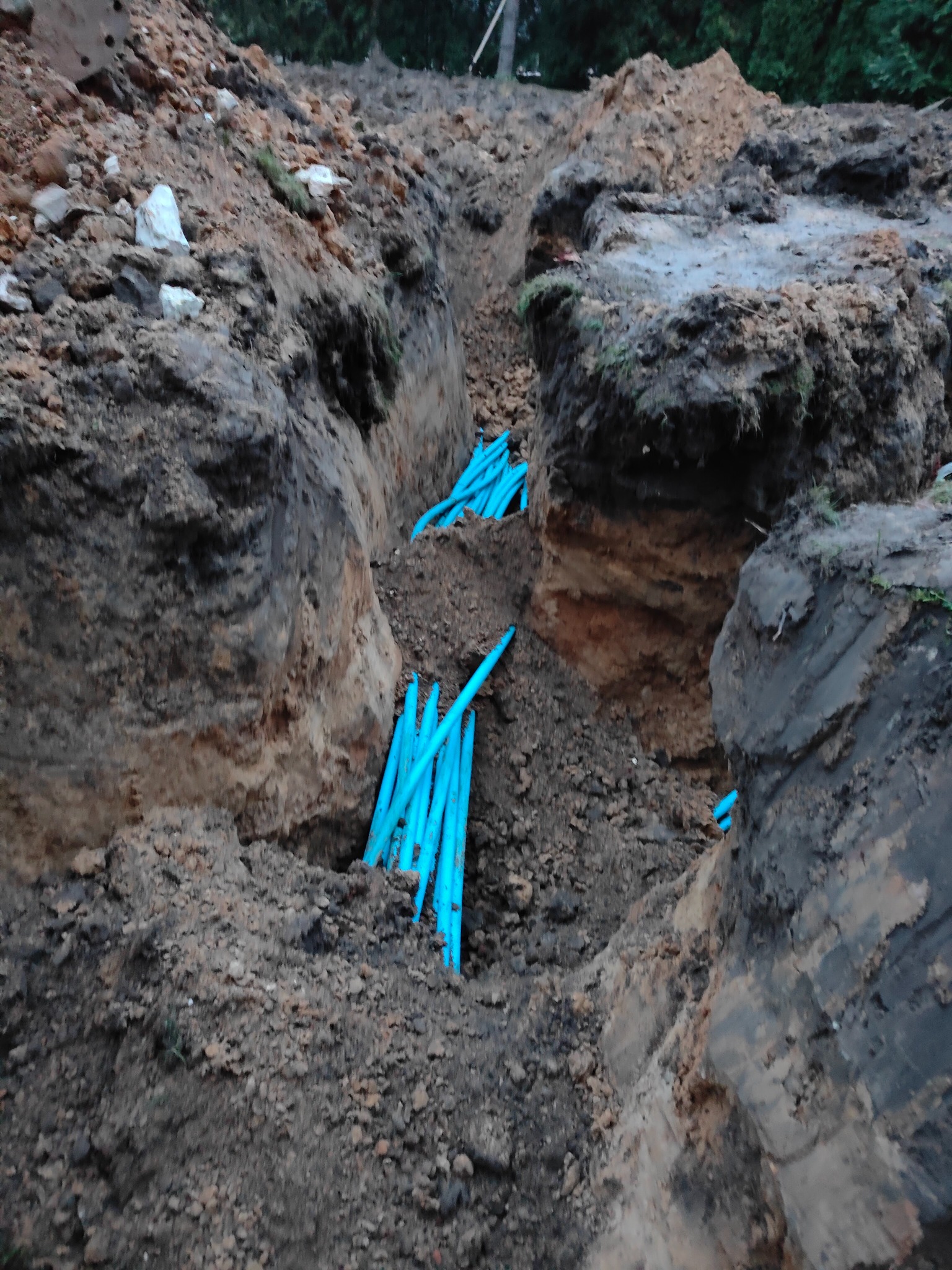 Wykop w ziemi z ułożonymi niebieskimi rurami, widoczne warstwy gleby i gruzu.